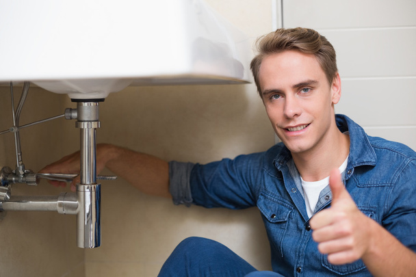 Handsome plumber gesturing thumbs up besides washbasin - Фото, изображение