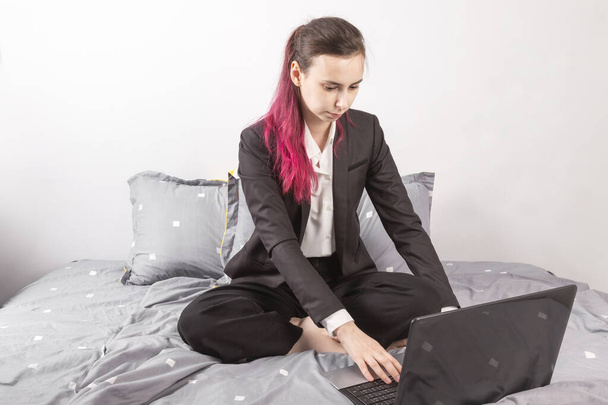 Konsepti - työtä kotoa, eristäytymistä, karanteenia. Nuori tyttö mustassa puvussa istuu sängyssä, pitäen kannettavaa polvillaan
 - Valokuva, kuva