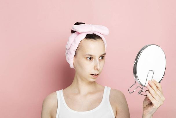 Young girl without makeup looks surprised in mirror on pink background - Foto, imagen