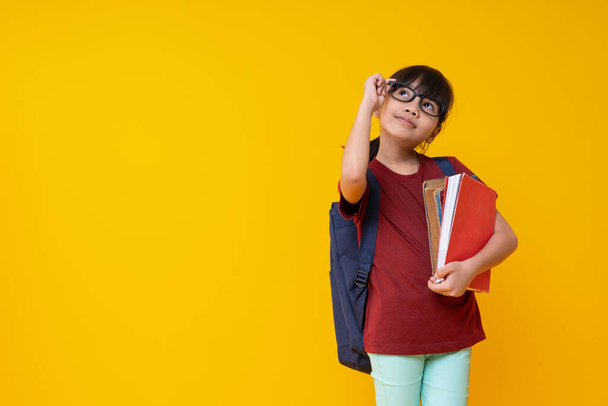 Portrait of Asian kid student holding book with glasses and looking up, Thai girl in red shirt thinking and get knowledge on yellow background in studio - Foto, Imagen