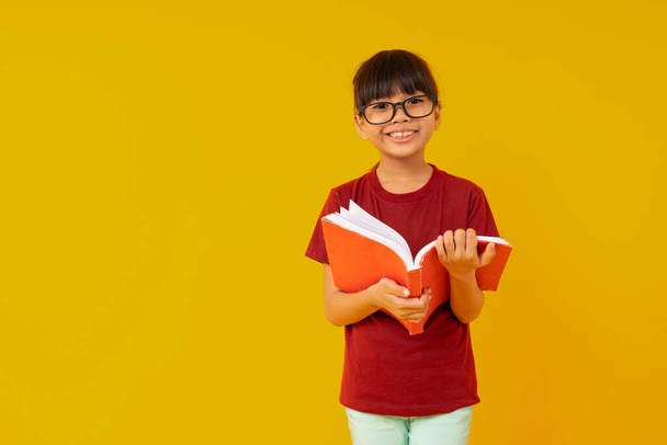 Jovem estudante da Ásia com grande sorriu vestindo óculos e camisa vermelha abrir e ler livro no fundo amarelo no estúdio  - Foto, Imagem