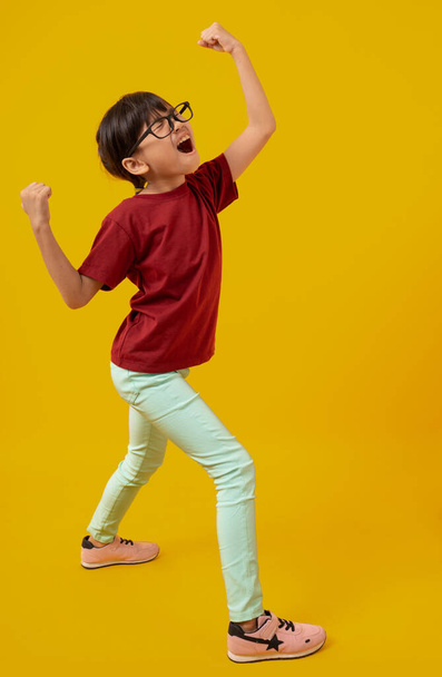 Portrait of Asian pretty girl celebrating and raising hand up for successful, Thai kid with glasses in red shirt have glad and joy on yellow in studio - Foto, Imagen