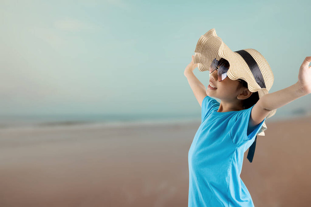 Joven niño bastante asiático con bolsa de viaje en estilo veraniego, chica tailandesa feliz con sombrero y gafas de sol mirando mar y playa en vacaciones
  - Foto, imagen