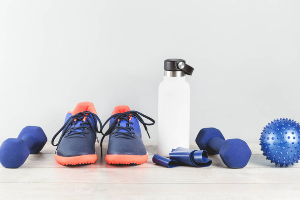 Healthy lifestyle blue and red gender neutral sports accessories grouped with white water bottle flask on white wood. Fitness and home or gym excersise monochrome items banner with copy space - Фото, изображение
