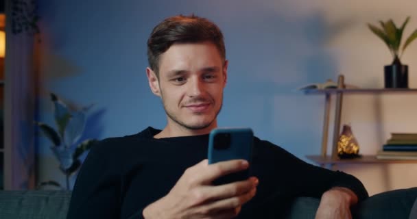 Excited brunnette good looking man using his smartphone while sitting on sofa at home. Close up view of handsome millennial guy looking and scrolling phone screen. Concept of leisure. - Video