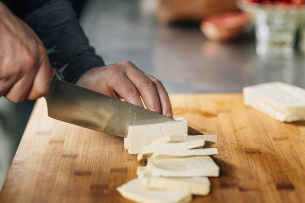 Preparing vegan meal. Chefs hand cutting tofu cheese with knife on wooden board  - Photo, Image
