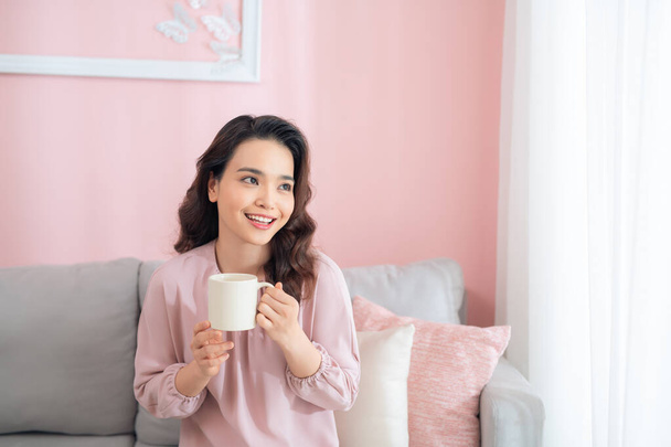 Cheerful young Asian woman holding a mug when sitting on sofa at home. - Фото, зображення