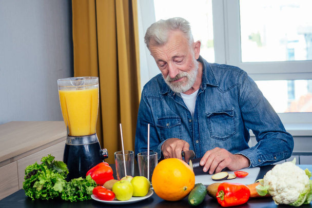 Elderly man making fresh vitamin juices smoothies and cut zucchini with a knife on the board bid window background - Foto, Imagem
