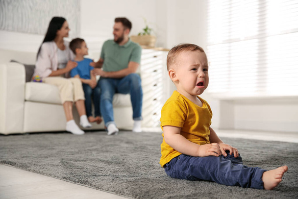 Unhappy baby sitting alone on floor while parents spending time with his elder brother at home. Jealousy in family - Zdjęcie, obraz