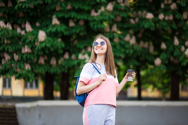 Take away coffee. Beautiful girl in sunglasses with backpack walk by summer street with coffee. Lifestyle concept - Foto, Imagem