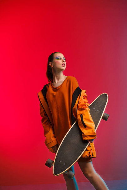 A young girl with bright makeup holds a longboard in her hands on a fashionable neon color background. Portrait of a girl skater - Fotografie, Obrázek
