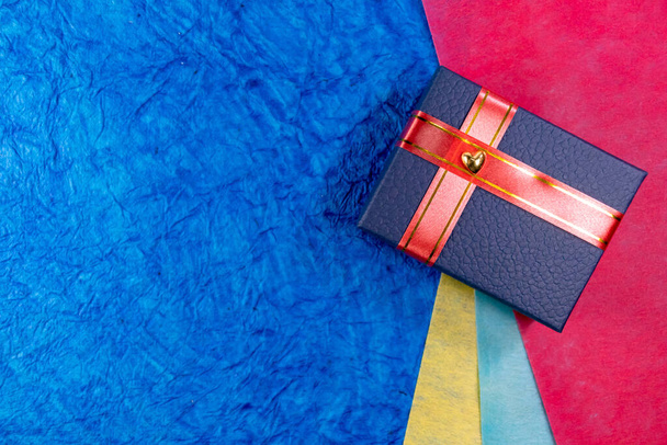 Close-up van een blauwe geschenkdoos met hart en rode linten over kleur papieren op de achtergrond. Begrip echtgenotes en echtgenoten geschenken - Foto, afbeelding