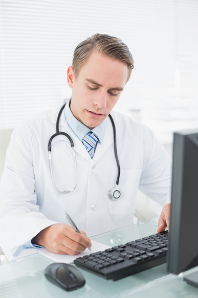 Doctor writing a note while using computer at medical office - Foto, afbeelding