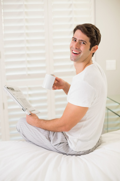 Cheerful man with coffee cup reading newspaper in bed - Foto, Imagem