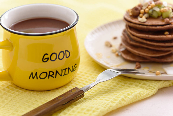 Nahaufnahme trendige Schokoladen-Pfannkuchen mit Haselnüssen, Kiwi, Kokosflocken und Ahornsirup in beigem Teller und gelber Tasse Kakao auf gelbem Küchentuch, Guten Morgen-Konzept - Foto, Bild