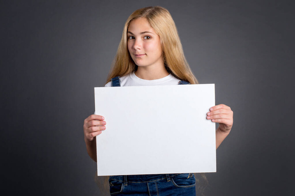 Χαριτωμένο έφηβο κορίτσι με μακριά ξανθά μαλλιά κρατώντας λευκό κενό αφίσα σε γκρι φόντο.   - Φωτογραφία, εικόνα