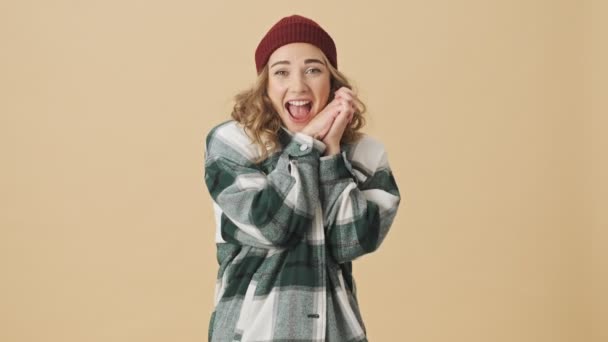 Pleased pretty woman in knit hat and shirt holding hands together and looking at the camera over beige background - Filmmaterial, Video