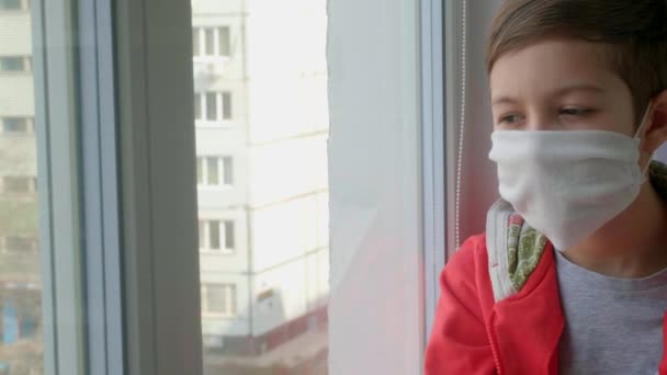 Un jeune garçon portant un masque médical regarde par la fenêtre. Auto-isolement en quarantaine, coronavirus, covide 19. - Séquence, vidéo