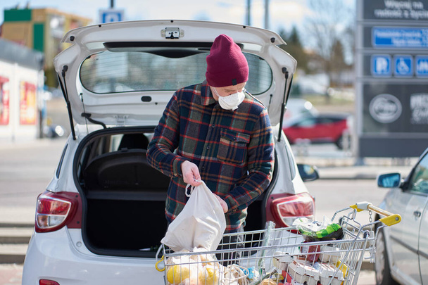 Caucasian man wearing medical mask packs bags with food from cart into car after shopping during outbreak - Photo, image