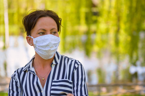 春の公園の女性は、彼女の後ろの湖の反射とコピースペースでパンデミックの間にコロナウイルスまたはCovid-19に対する感染制御として顔のマスクを身に着けている - 写真・画像