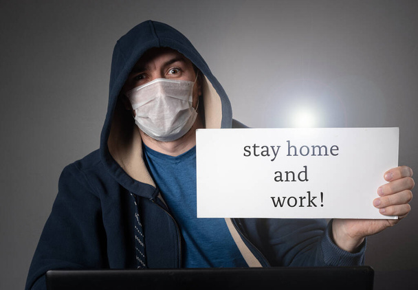 male hacker in a medical mask and a hood, works at a laptop and holds a tablet with the inscription - stay home and work - Фото, изображение