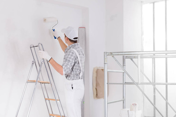 Malarz stojący na drabinie malujący białą ścianę w pomieszczeniu w trakcie budowy odchylający się za pomocą rolki oglądanej od tyłu z ramą rusztowania wewnętrznego obok - Zdjęcie, obraz