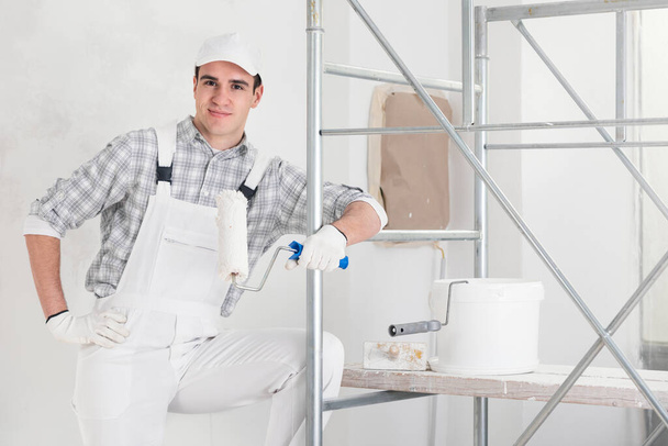 Confident relaxed young painter at work leaning on mobile interior scaffolding holding a roller with tub of paint smiling at the camera in a DIY, renovation or construction concept - 写真・画像