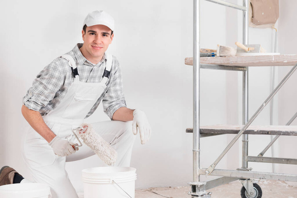 Friendly young painter or decorator in overalls and white cap smiling at the camera with a paint roller in his hand in a DIY, renovation or construction concept - Photo, Image