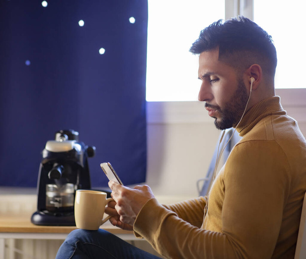 Un hombre guapo en la cocina está sonriendo, tomando café por la mañana y mirando el teléfono inteligente con auriculares
. - Foto, imagen