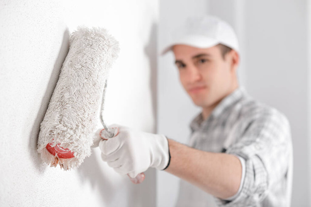 Painter painting a white wall with a roller with selective focus to the roller and his hand in the foreground and copy space in a DIY, renovation or construction concept - Photo, Image