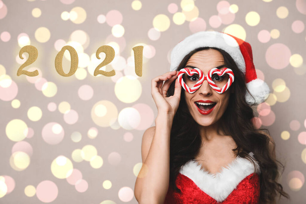 Emotional young woman in Christmas costume with party glasses on beige background. Bokeh effect - Foto, imagen
