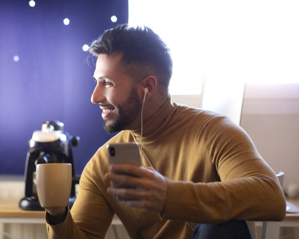 Красивый мужчина на кухне улыбается, пьет утренний кофе и смотрит на смартфон в наушниках
. - Фото, изображение
