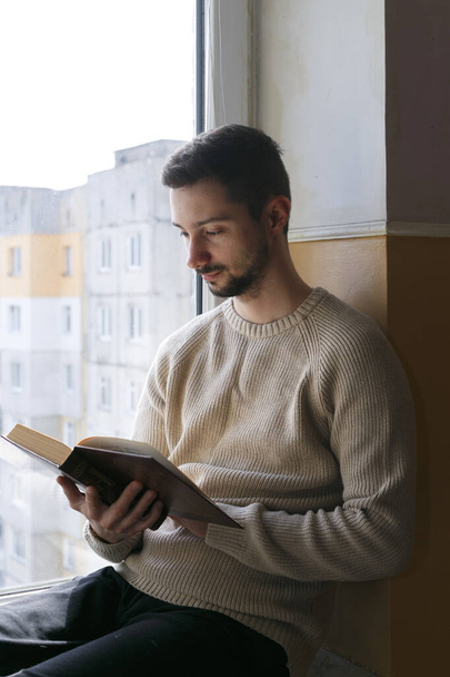 Ein junger Mann sitzt auf einer Fensterbank und liest ein Buch. Ein Mann trägt einen beigen Pullover. Der Mann ist bärtig. - Foto, Bild