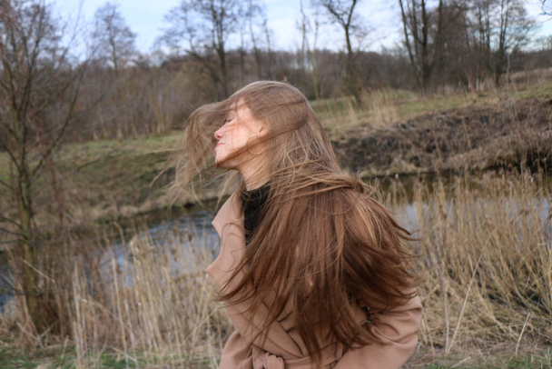 piękna dziewczyna, blondynka z długimi włosami, spaceruje wiosną w pobliżu lasu w brązowym płaszczu., spokój i wolność - Zdjęcie, obraz