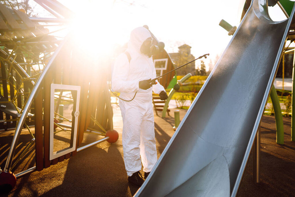 Korunaklı kıyafet giyen bir adam karantina şehrinde koronavirüsün yayılmasını önlemek için güneş altındaki oyun alanını sprey kimyasallarla dezenfekte ediyor. Covid -19. Temizlik kavramı. - Fotoğraf, Görsel