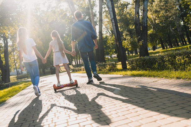 Rear view of young parents on sunny day walking in park with their cute little girl riding hoveboard - Fotoğraf, Görsel