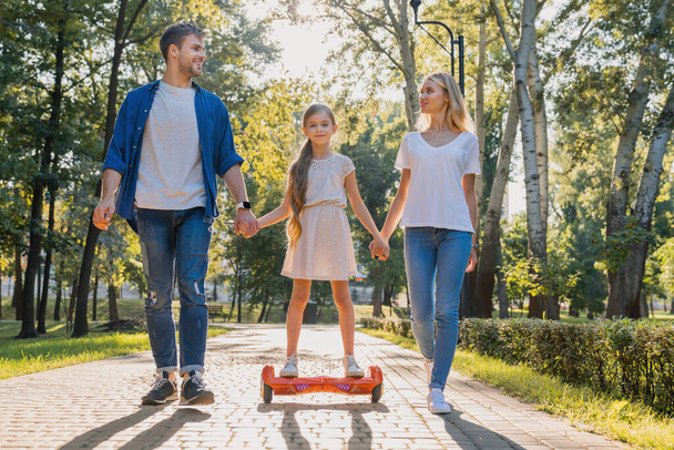 Young parents walking in park with their cute little girl riding hoveboard - Foto, afbeelding