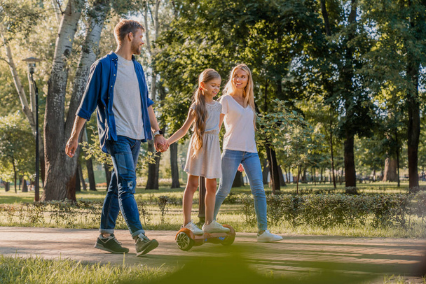 Vista lateral de los padres sonrientes jóvenes caminando en el parque con su linda niña montando hoveboard
 - Foto, imagen