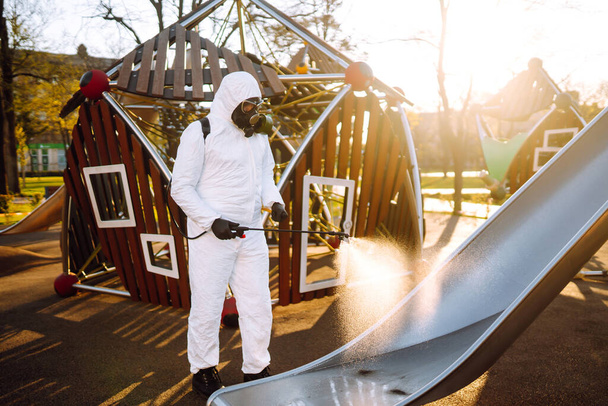 Человек в защитном костюме дезинфицирует детскую площадку на солнце с помощью химикатов для предотвращения распространения коронавируса, пандемии в карантинном городе. Ковид - 19. Концепция очистки
. - Фото, изображение