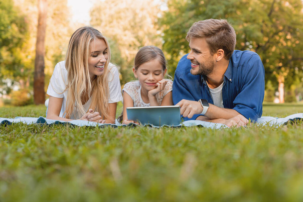 Güzel, genç bir aile piknik battaniyesinde uzanıp dijital tablet kullanırken sonbahar gününün tadını çıkarıyor. - Fotoğraf, Görsel