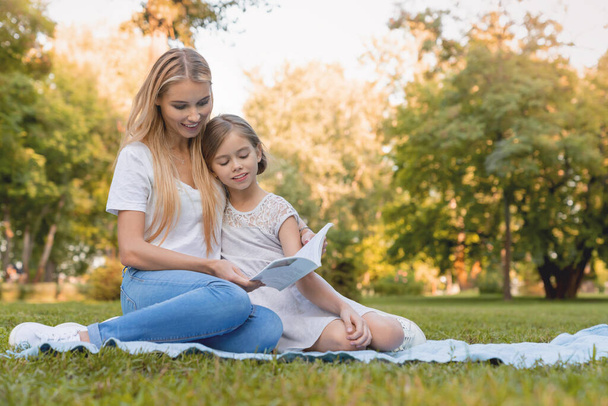 Mother with her cute little daughter reading book in the park - Zdjęcie, obraz