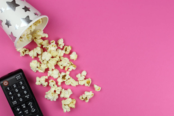 TV of DVD afstandsbediening en een omgekeerde Beker met gemorste popcorn op een roze achtergrond - Foto, afbeelding