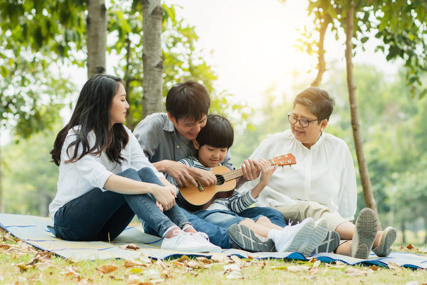 Familia feliz con la abuela, mamá con papá enseñando a tocar la guitarra hijo y cantar una canción en el parque, Disfrutar y relajarse gente picnic al aire libre
 - Foto, imagen