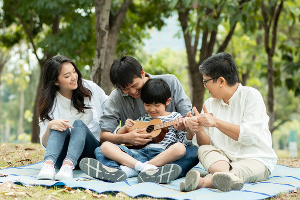 Ευτυχισμένη οικογένεια με τη γιαγιά, μαμά με τον μπαμπά διδασκαλία γιο παίζει κιθάρα και να τραγουδήσει ένα τραγούδι στο πάρκο, Απολαύστε και χαλαρώστε τους ανθρώπους πικνίκ έξω  - Φωτογραφία, εικόνα