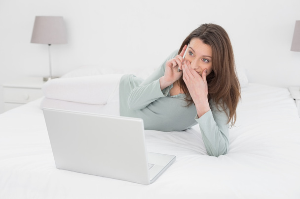Shocked casual woman using cellphone and laptop in bed - Photo, image