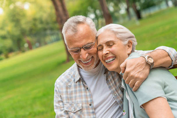 Gülümseyen adam ve kadın parkta bankta oturuyor sarılıyor ve gülüyorlar. - Fotoğraf, Görsel