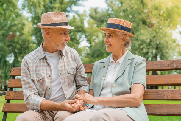 Front view of happy senior couple in hats holding hands and looking on each other with smile while sitting on the park bench together - Fotografie, Obrázek