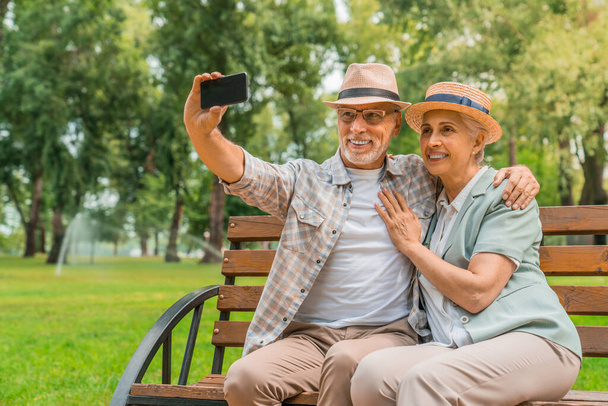 Старшая пара в шляпах обнимает и делает селфи на мобильном телефоне, сидя на скамейке на открытом воздухе
 - Фото, изображение