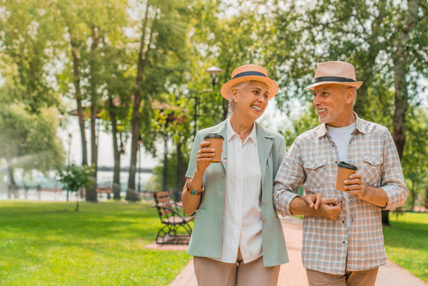 Happy elderly man and woman walking in park and drinking coffee - Фото, изображение