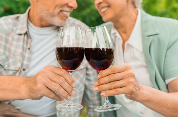 Cropped image of beautiful senior couple drinking wine and enjoying time together while sitting outdoor, focus on glasses - Fotografie, Obrázek